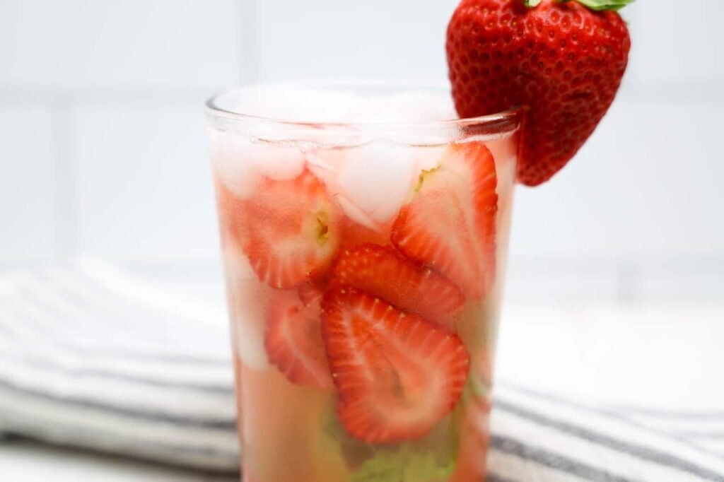 Close-up glass of strawberry basil lemonade filled with sliced strawberries and ice with a fresh strawberry garnish on the rim.