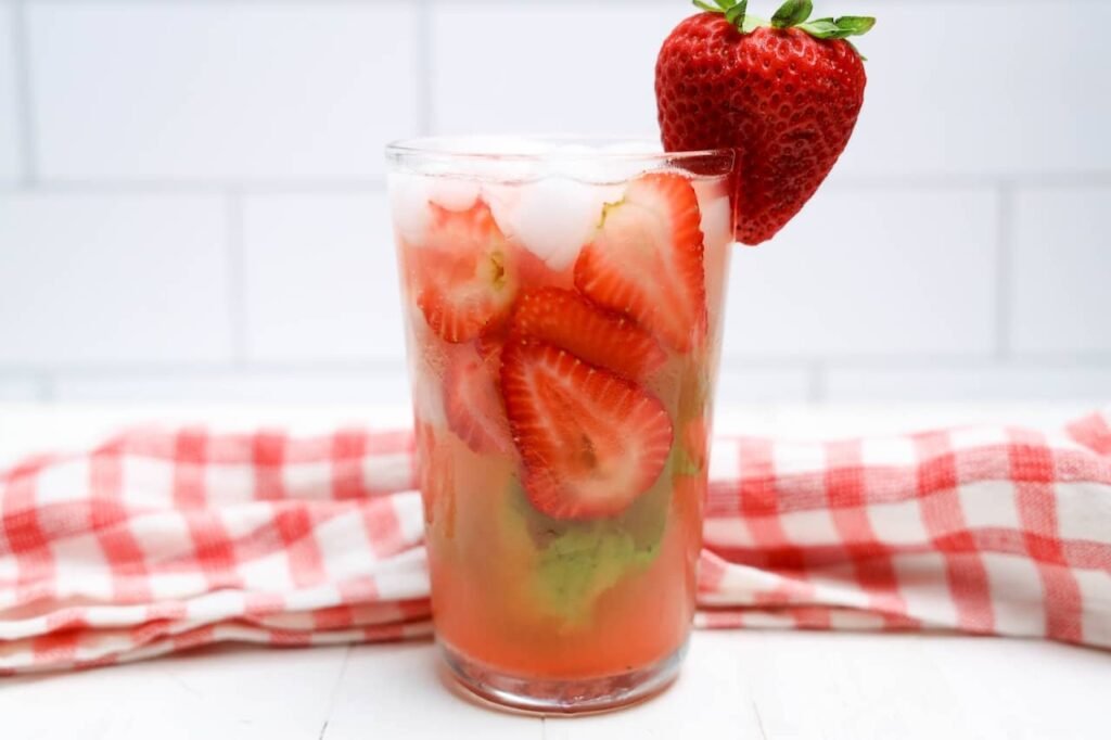 Glass of strawberry basil lemonade filled with sliced strawberries, basil leaves, and ice with a fresh strawberry garnish and red gingham cloth in the background.