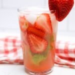 Glass of strawberry basil lemonade filled with sliced strawberries, basil leaves, and ice with a fresh strawberry garnish on the rim.