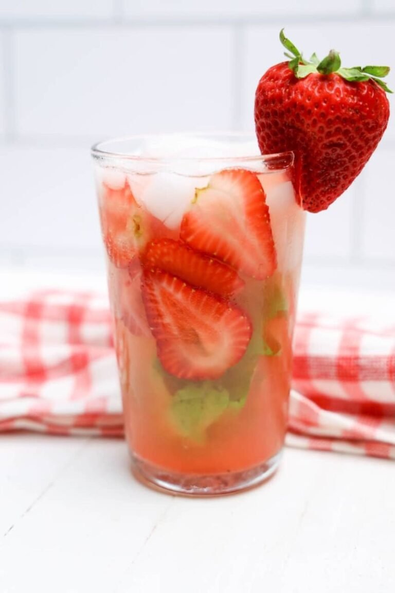 Glass of strawberry basil lemonade filled with sliced strawberries, basil leaves, and ice with a fresh strawberry garnish on the rim.