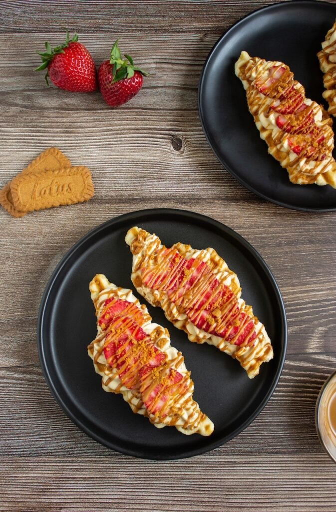 Two finished Biscoff strawberry croffles served on black plates and topped with piped Biscoff whipped cream, fresh sliced strawberries, melted Biscoff drizzle, and crushed Biscoff cookies. The croffles have a crispy golden exterior with creamy, fruity, and cookie butter toppings, ready to serve as an easy brunch or dessert.